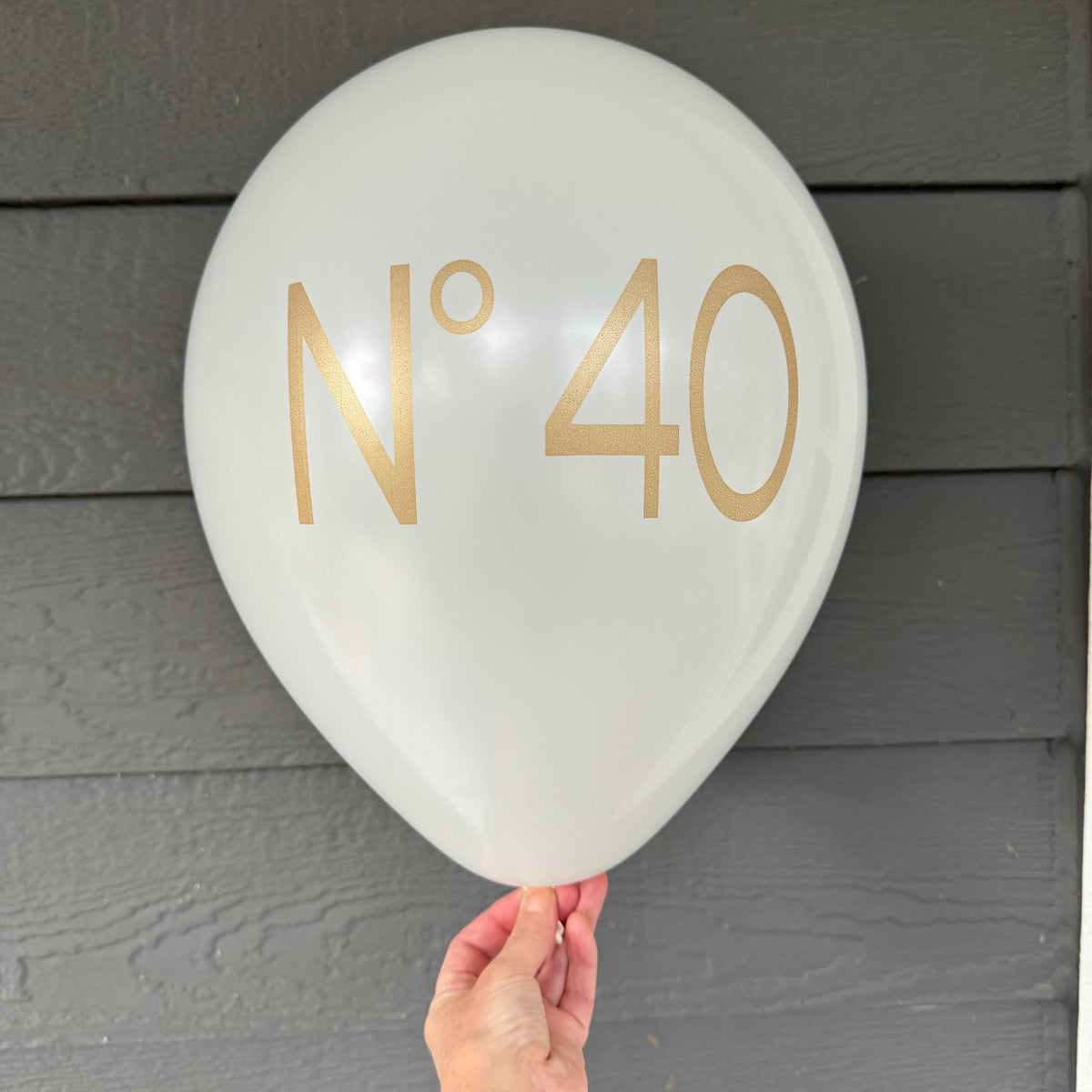 Hand holding a white No 40 birthday balloon with gold lettering against gray siding.