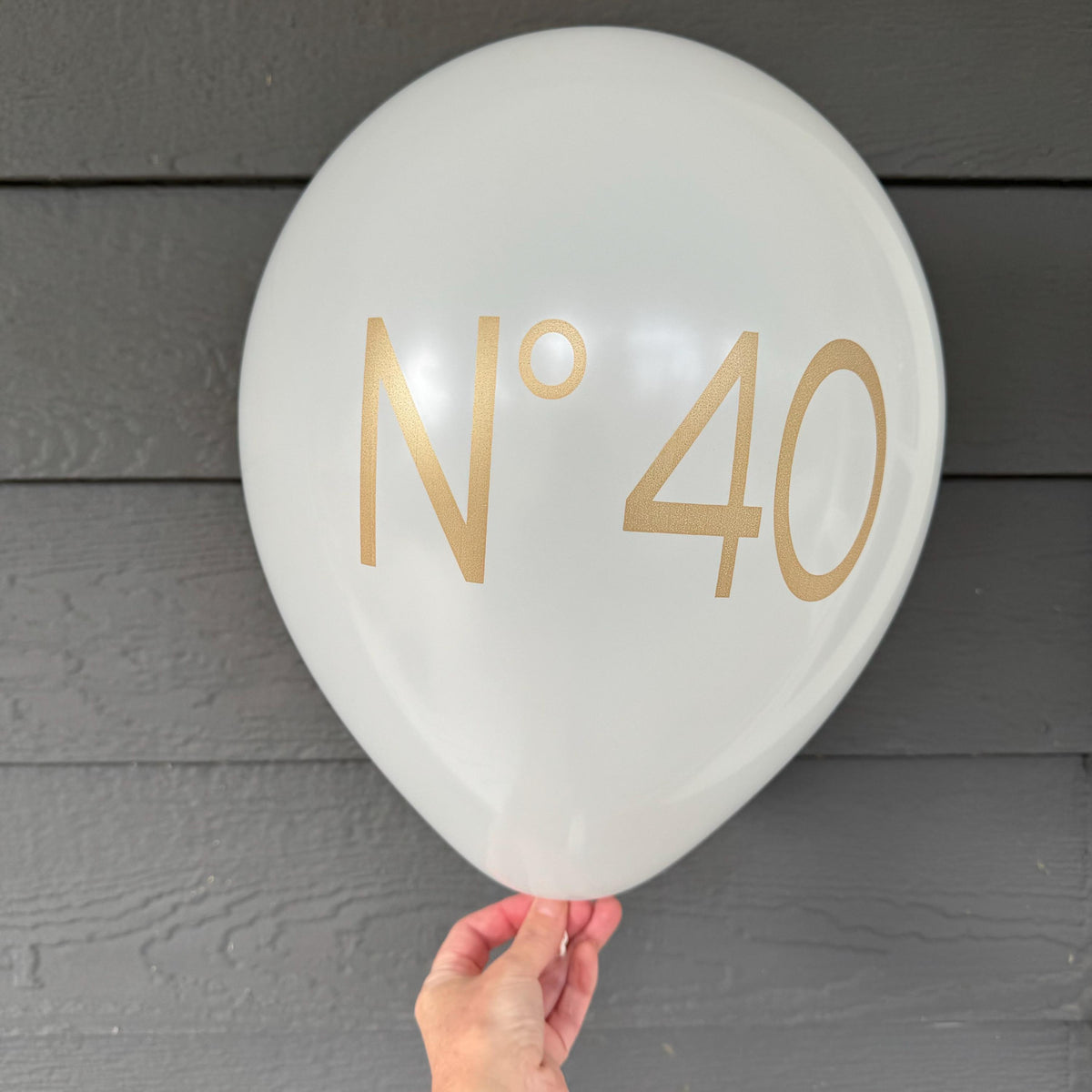 White No 40 latex birthday balloon with gold lettering, held in front of gray horizontal siding.