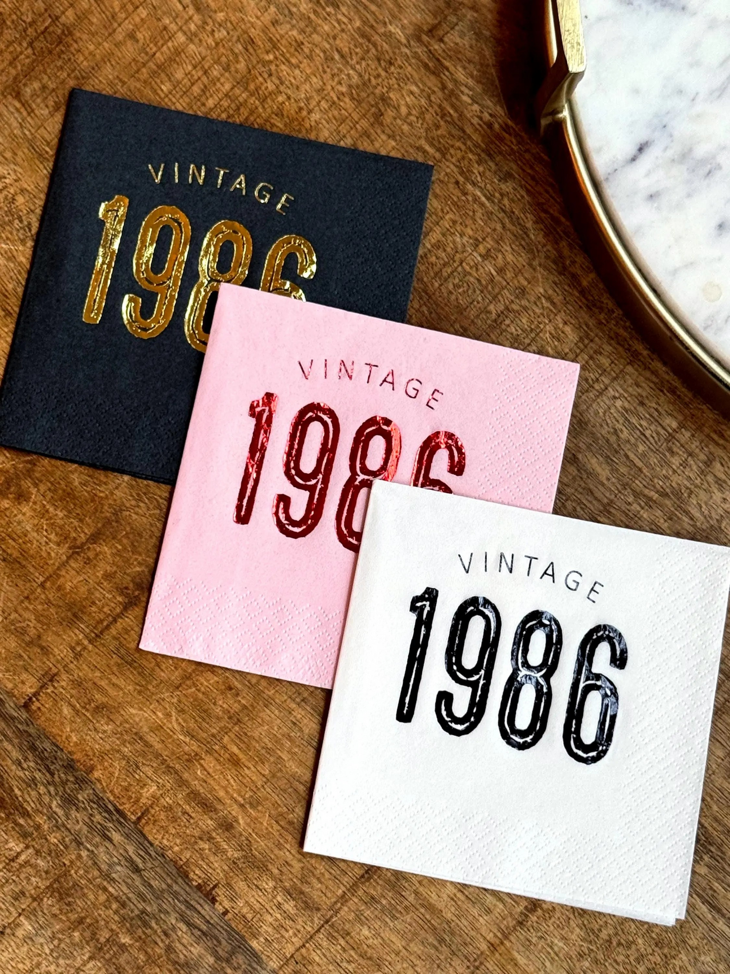 Three 'Vintage 1986' cocktail napkins in black, pink, and white on a wooden surface.