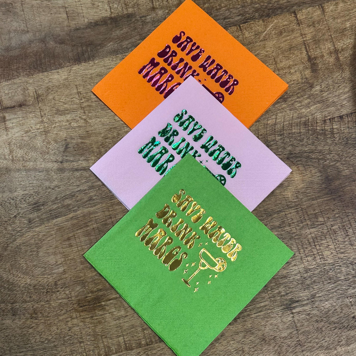 Three folded cocktail napkins in orange, pink, and lime green are stacked on a wooden surface with a foil-stamped cocktail design.