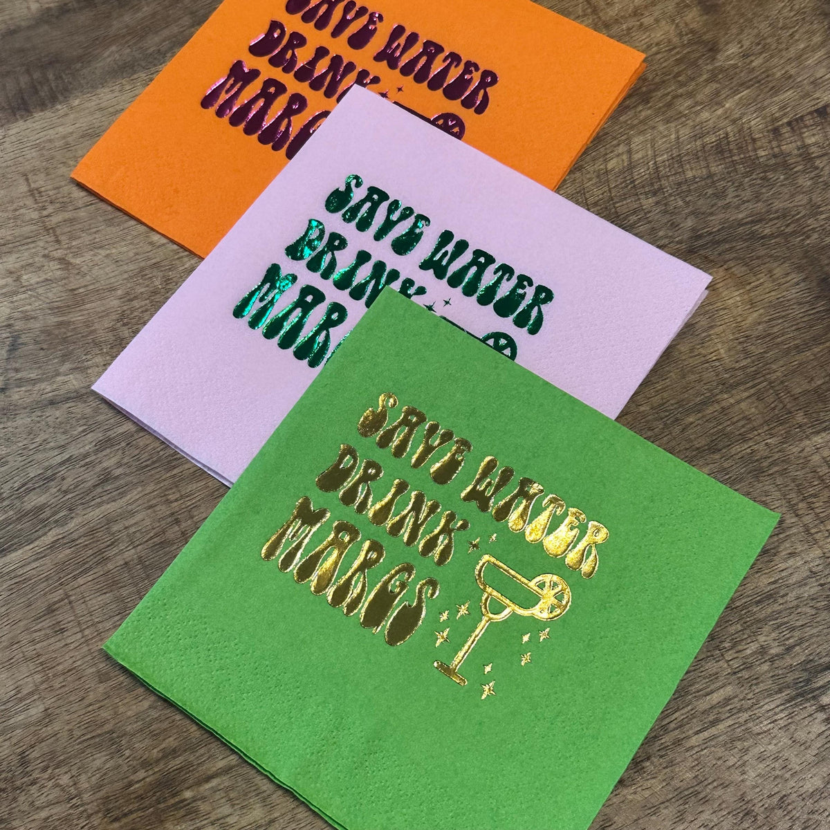 Three folded paper cocktail napkins stacked diagonally on a wood surface, featuring orange with purple foil text, light pink with green foil text, and green with gold foil text reading save water drink margs.