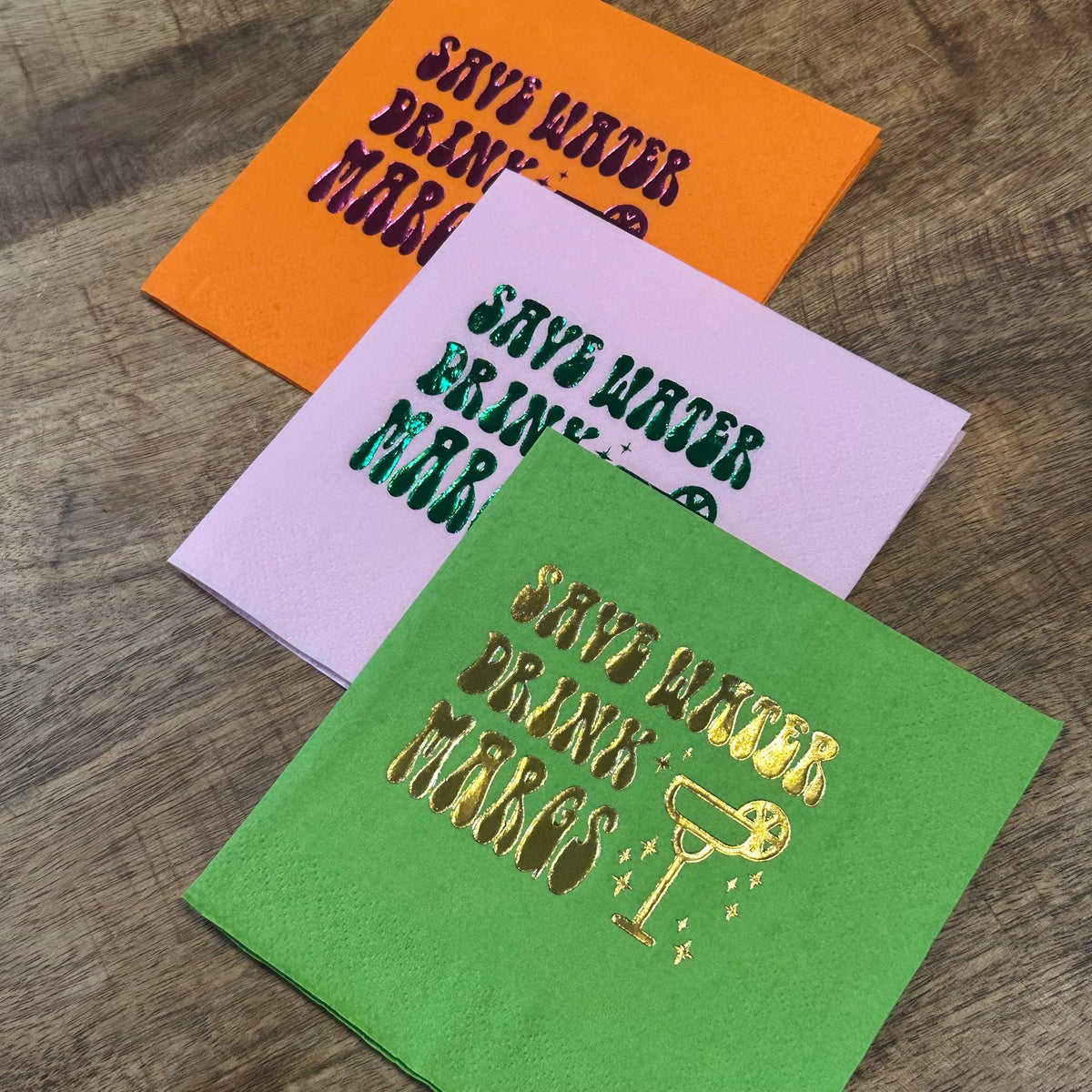 A close-up of three colorful folded cocktail napkins on a wooden background, showing an orange napkin with purple foil text, a pink napkin with green foil text, and a green napkin with gold foil text.