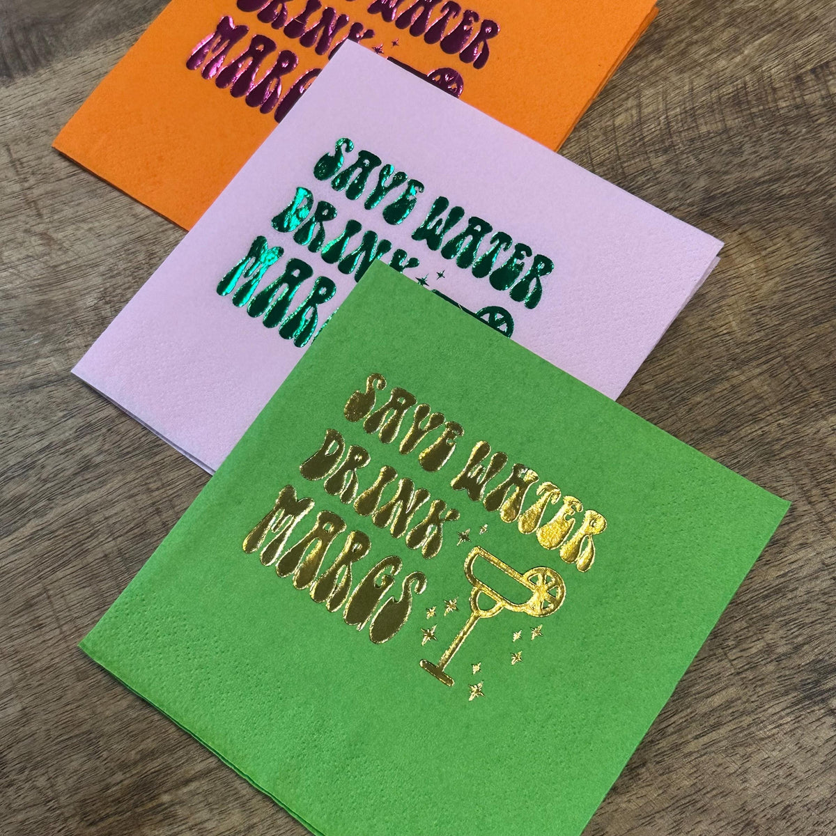 An angled shot of three stacked cocktail napkins on a rustic wooden table, with an orange, a light pink, and a bright green napkin displaying shiny foil lettering.