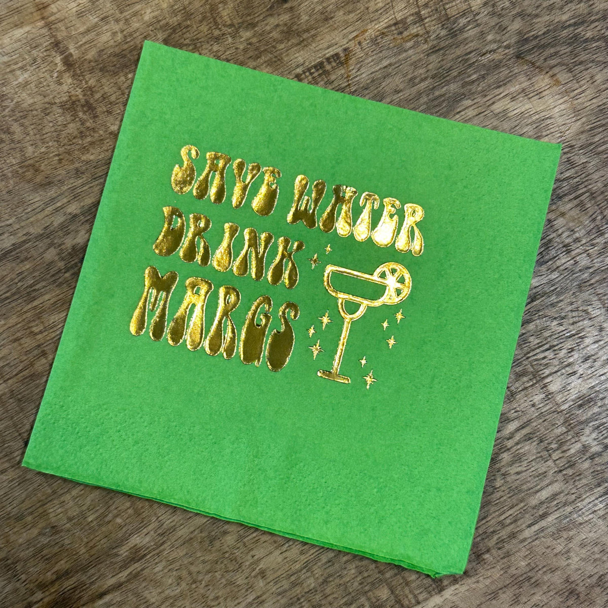 A single bright green folded cocktail napkin resting on a wood grain surface, printed with metallic gold foil text and a margarita glass graphic.