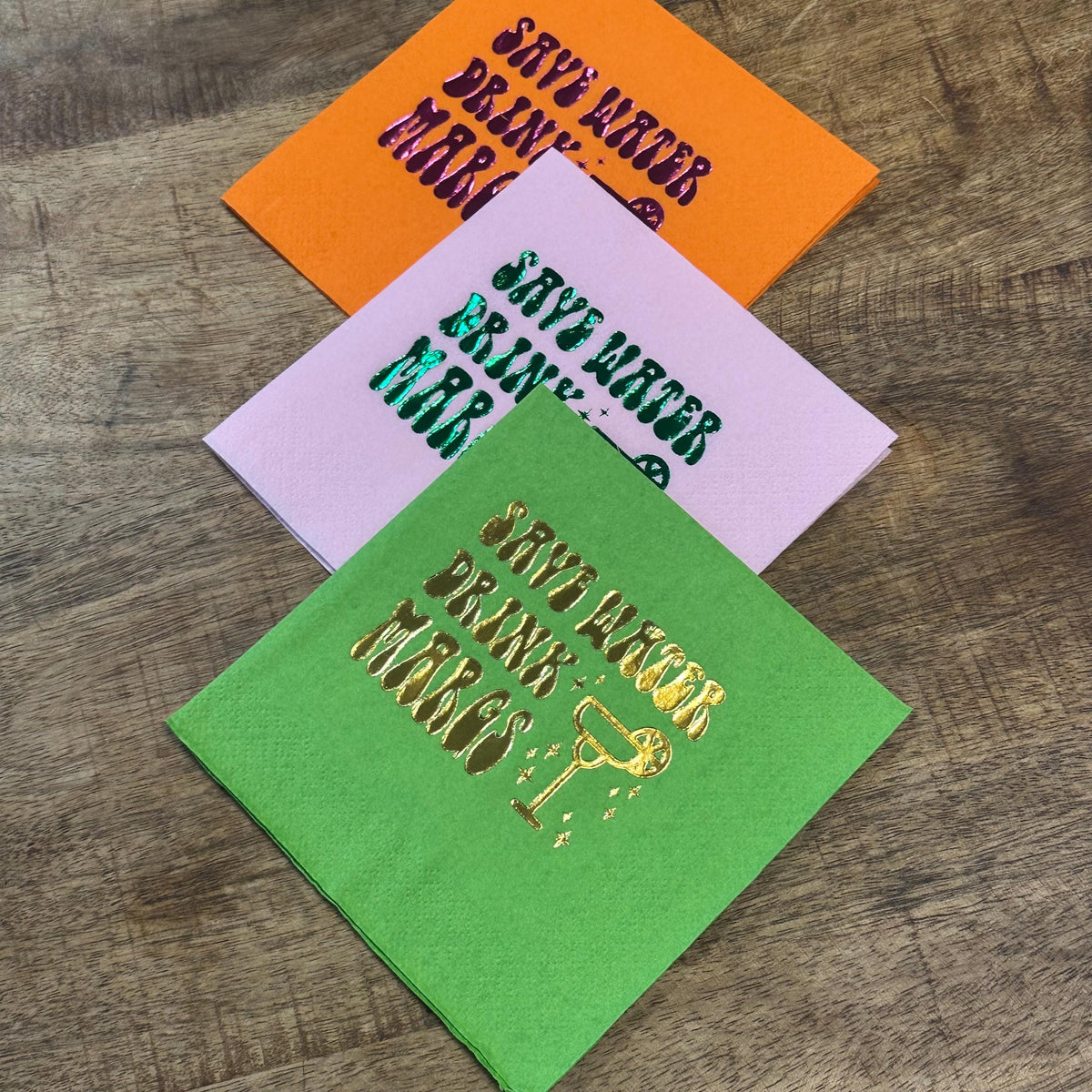 Three folded cocktail napkins arranged in a diagonal stack on a wooden surface, displaying orange, light pink, and green colors with shiny foil printed text.