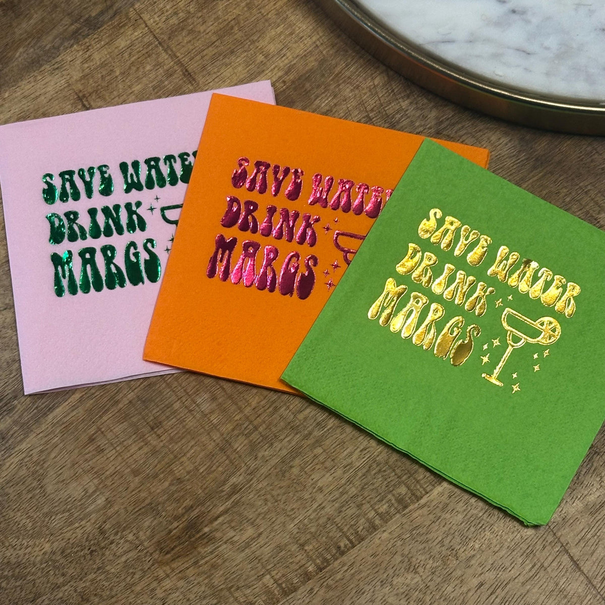 Three folded cocktail napkins fanned out on a wooden tabletop, including a pink napkin with dark green foil, an orange napkin with magenta foil, and a green napkin with gold foil.