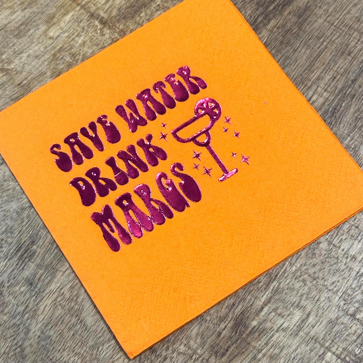An orange cocktail napkin printed with shiny purple foil text that reads Save Water Drink Margs next to a margarita glass illustration.