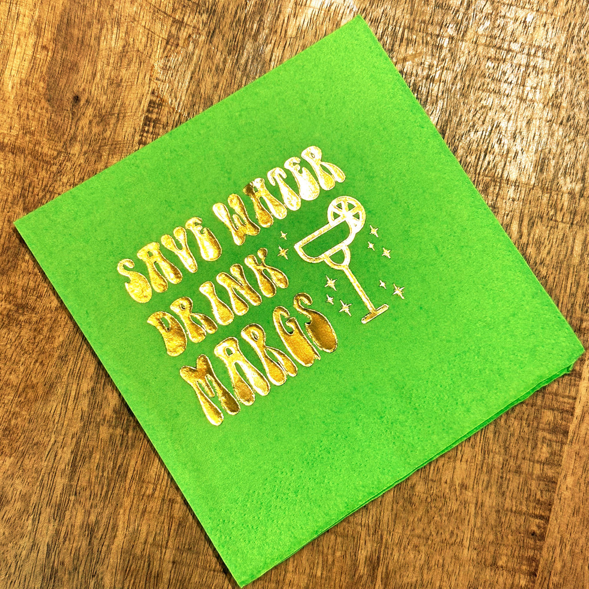 A lime green cocktail napkin featuring metallic gold foil text reading Save Water Drink Margs with a margarita glass graphic.
