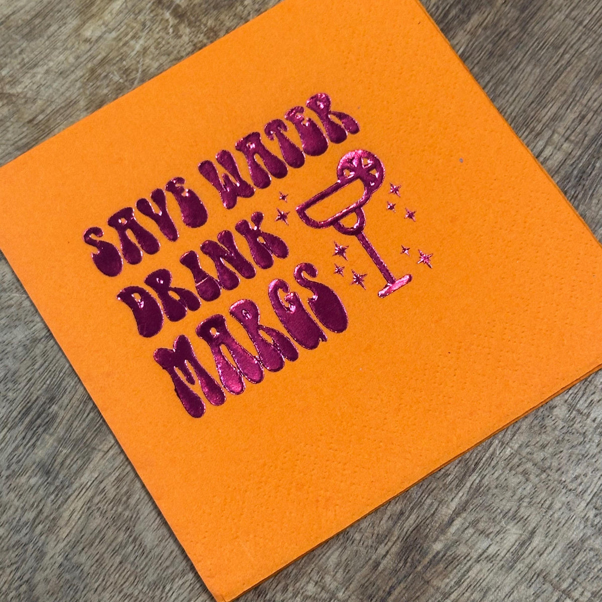 An orange cocktail napkin featuring metallic pink foil lettering that reads Save Water Drink Margs alongside a margarita glass icon.