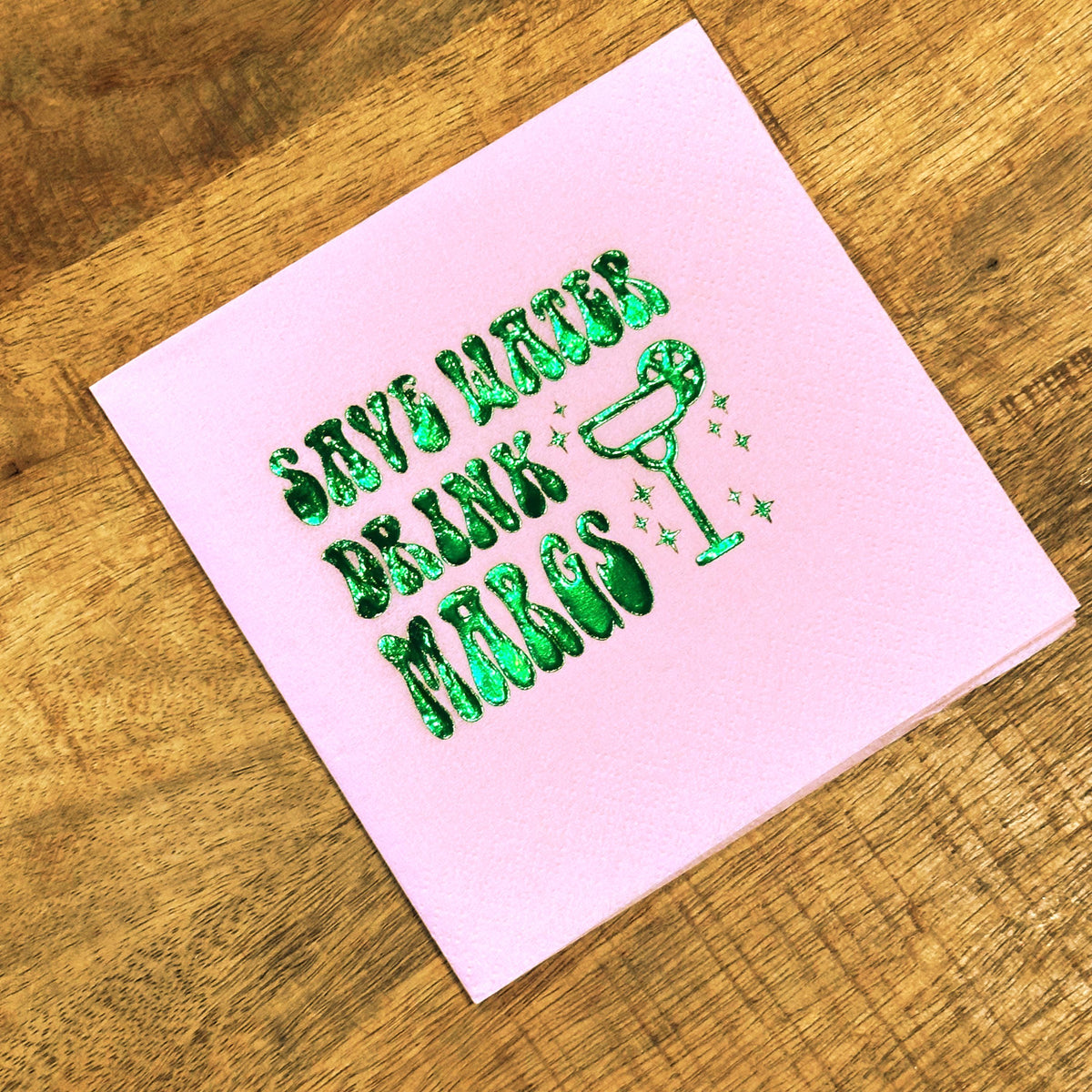 A pastel pink cocktail napkin with shiny green foil text that says Save Water Drink Margs beside a margarita glass outline.