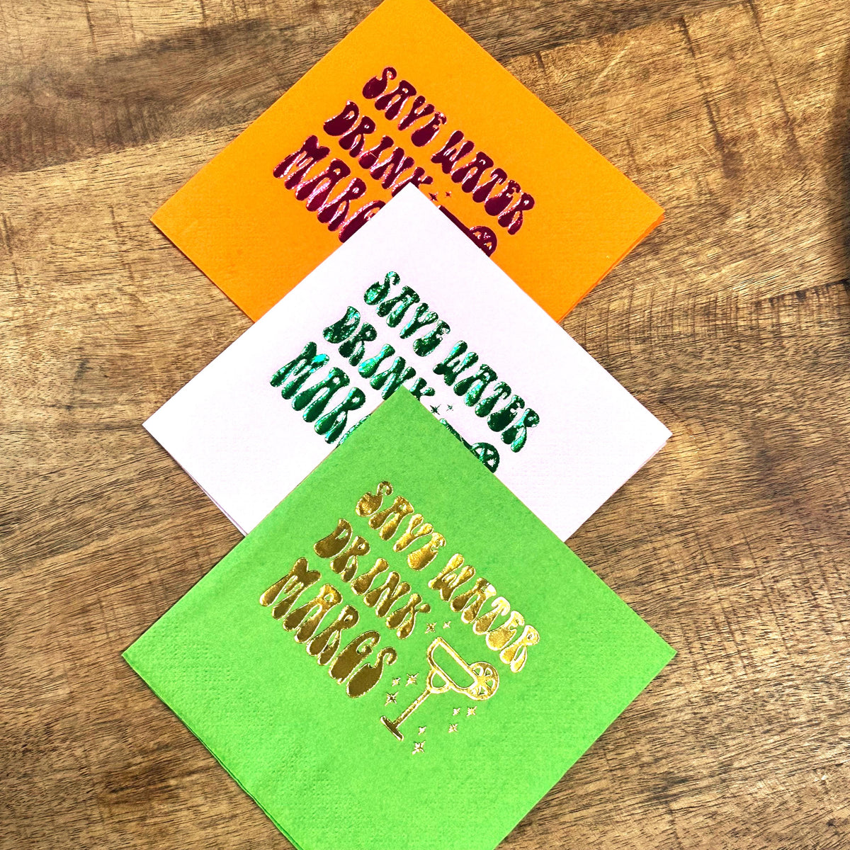 Three folded cocktail napkins stacked diagonally on a wood surface in orange, white, and lime green with varying foil colors.