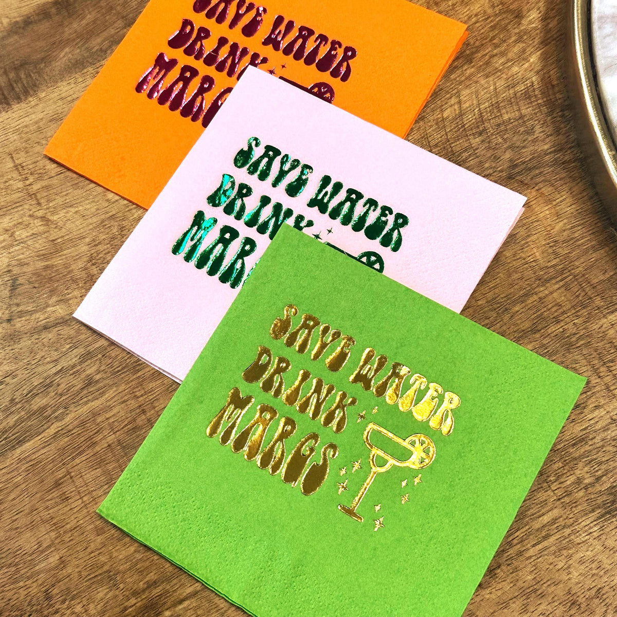 Three stacked cocktail napkins in orange, pink, and green featuring foil-stamped text that reads Save Water Drink Margs.