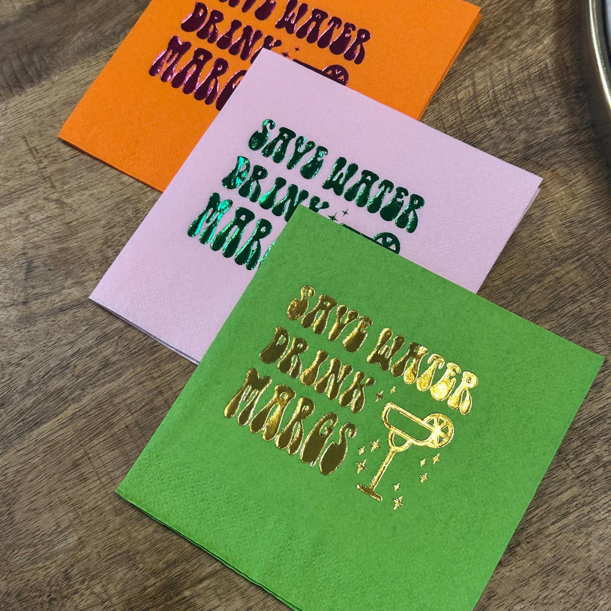 Three layered cocktail napkins in bright orange, light pink, and lime green with shiny foil text and a margarita glass design.