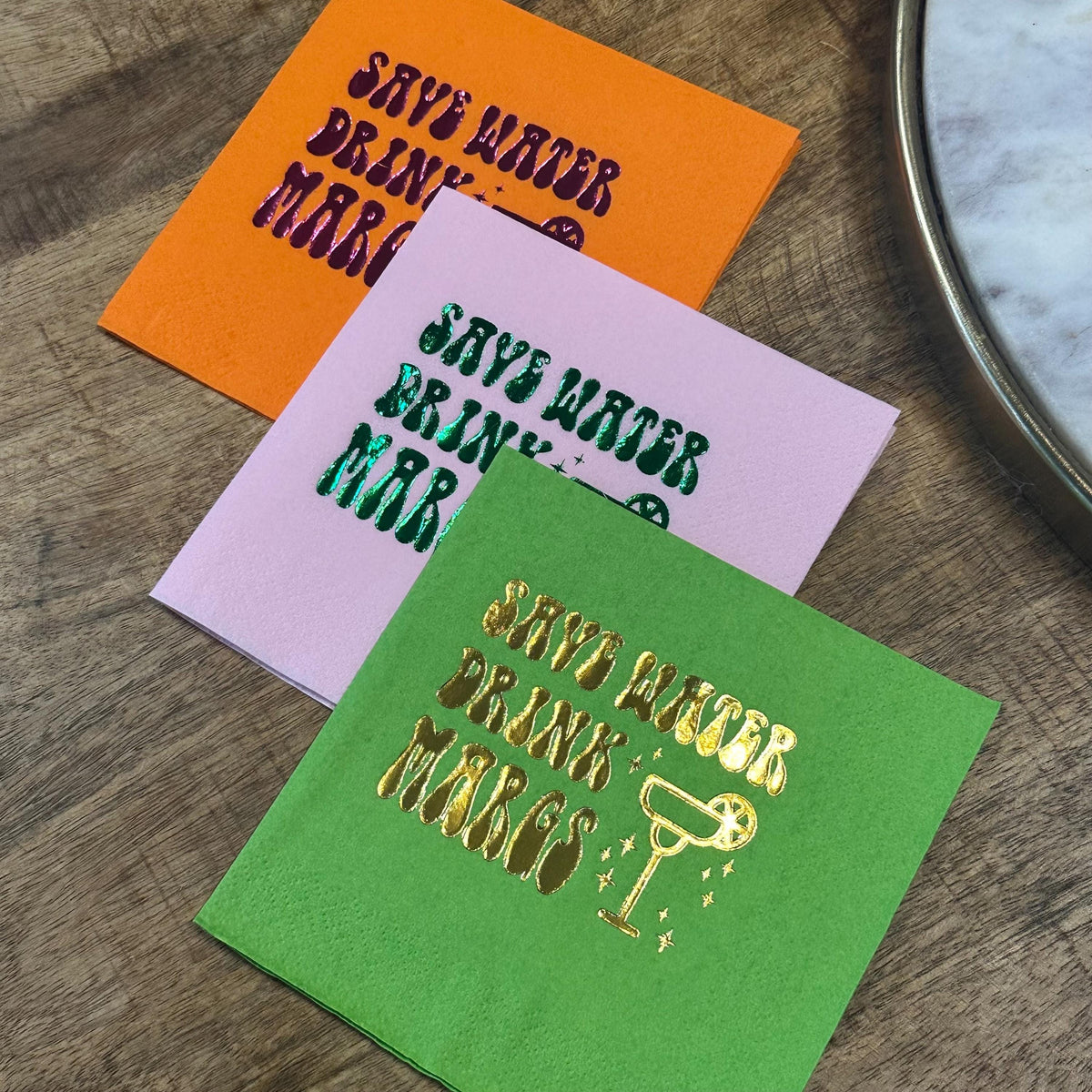 A flatlay of three stacked cocktail napkins in orange, pink, and green placed next to a circular tray on a wooden table.