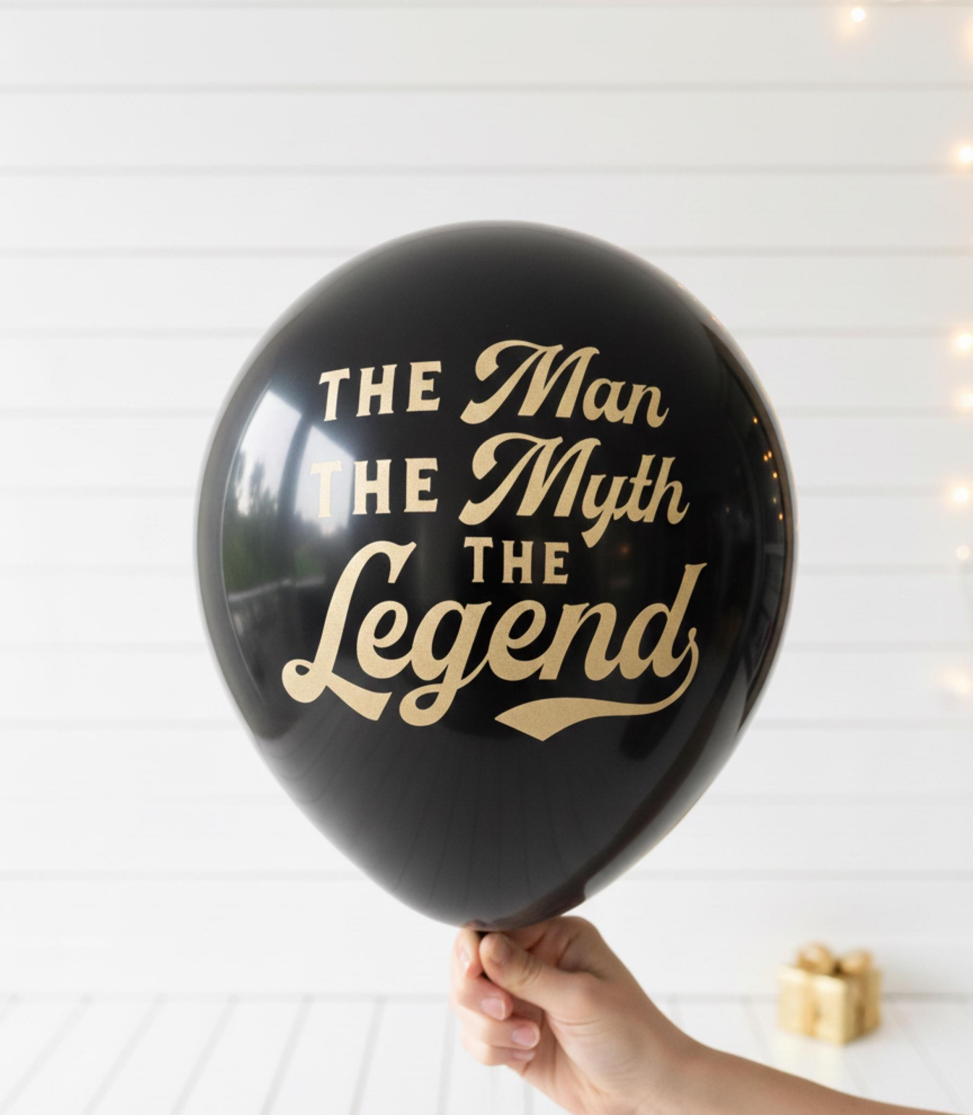 Black balloon with gold text 'The Man, The Myth, The Legend' held by a hand against a white background.  The font is in gold 