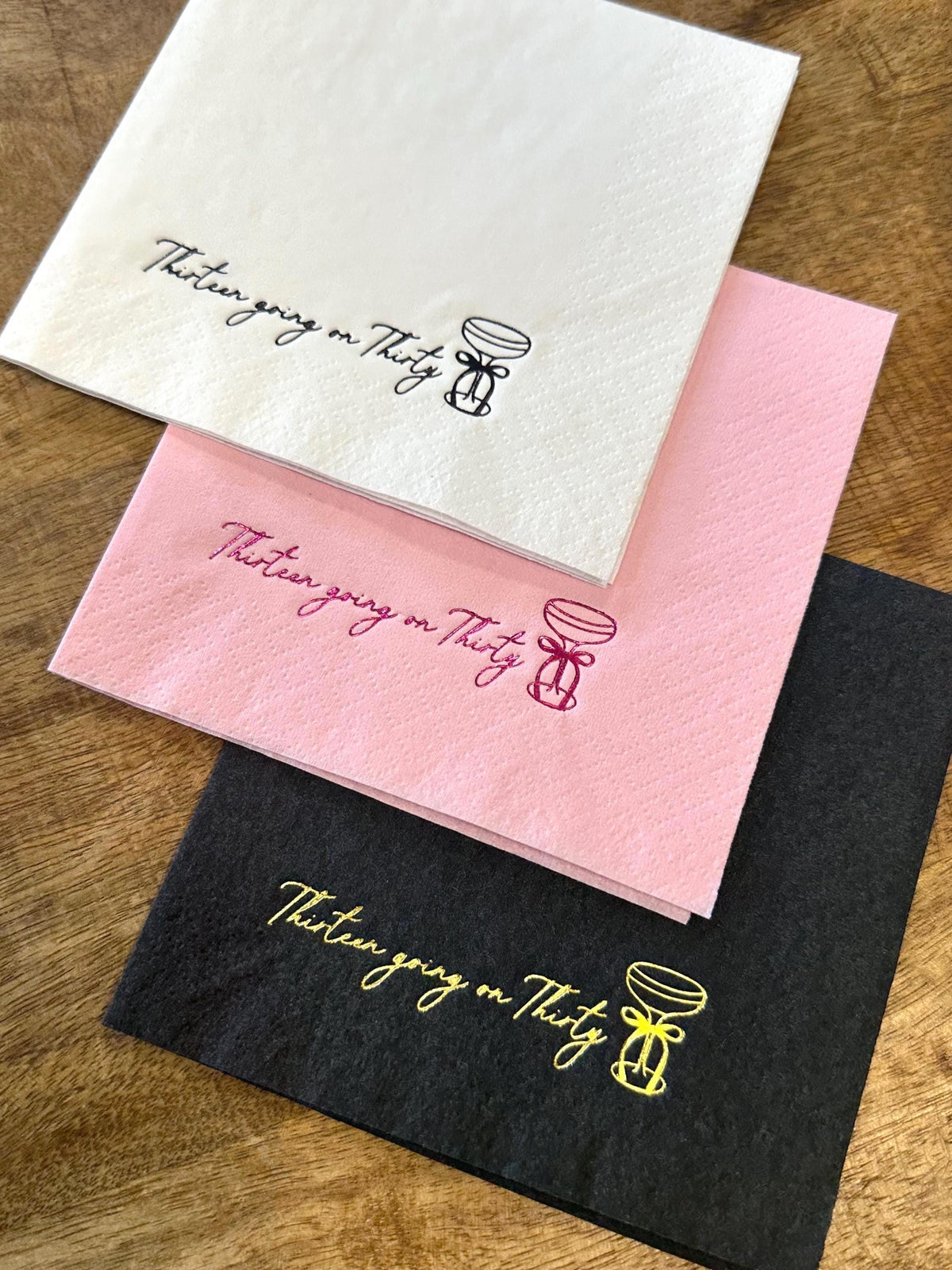 Three folded paper napkins in white, pink, and black stacked on a wooden surface, each featuring a foil-stamped cocktail glass and the text Thirteen going on Thirty.