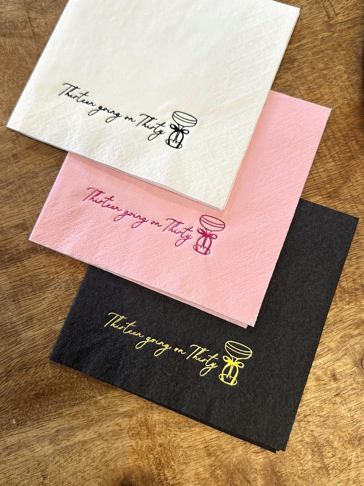 Three folded paper napkins in white, pink, and black overlapping on a wooden surface, each stamped with a cocktail glass and the text Thirteen going on Thirty.
