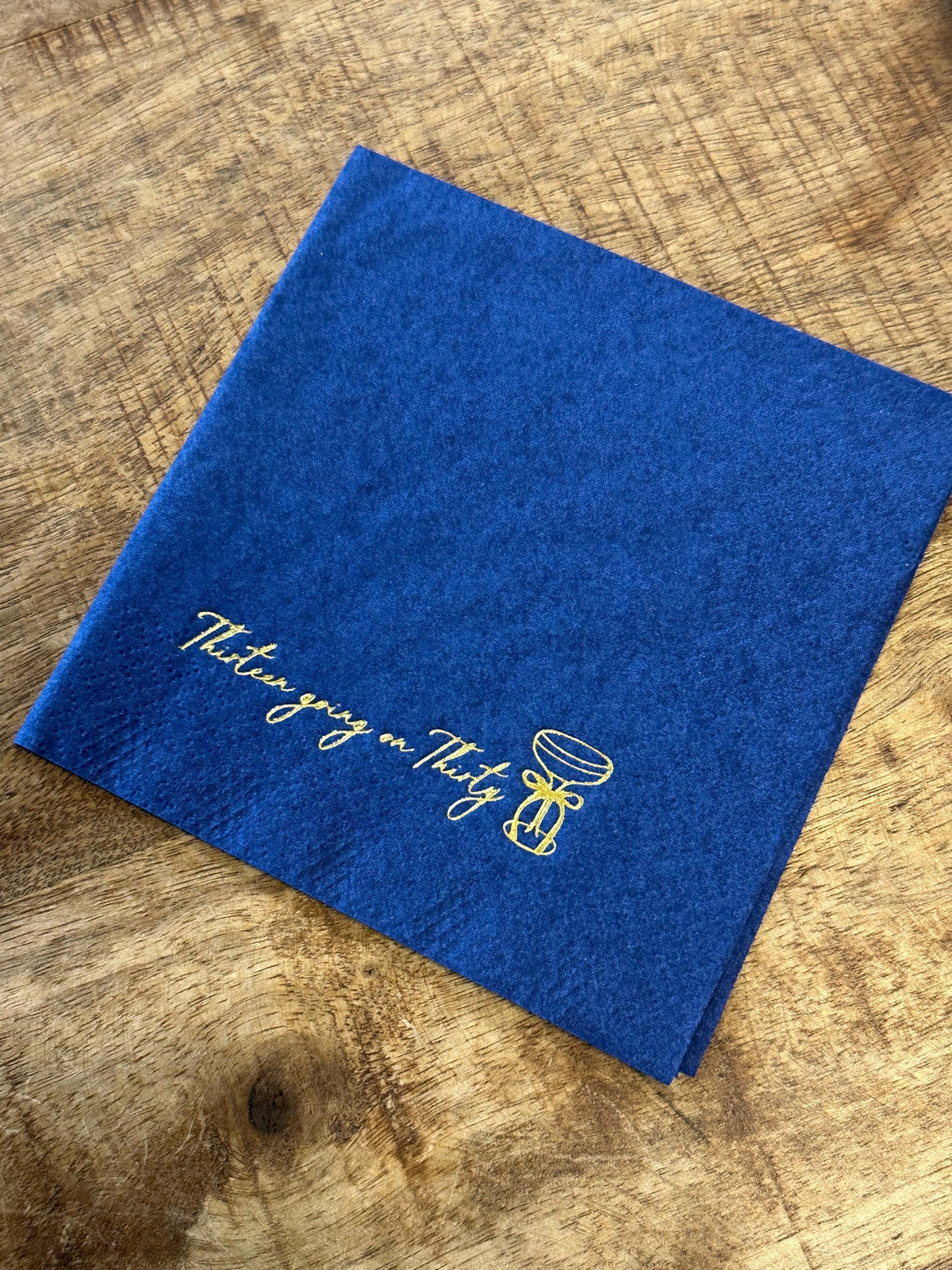 A square blue paper napkin lies flat on a wooden surface, featuring gold foil text that reads Thirteen going on Thirty with a martini glass graphic.