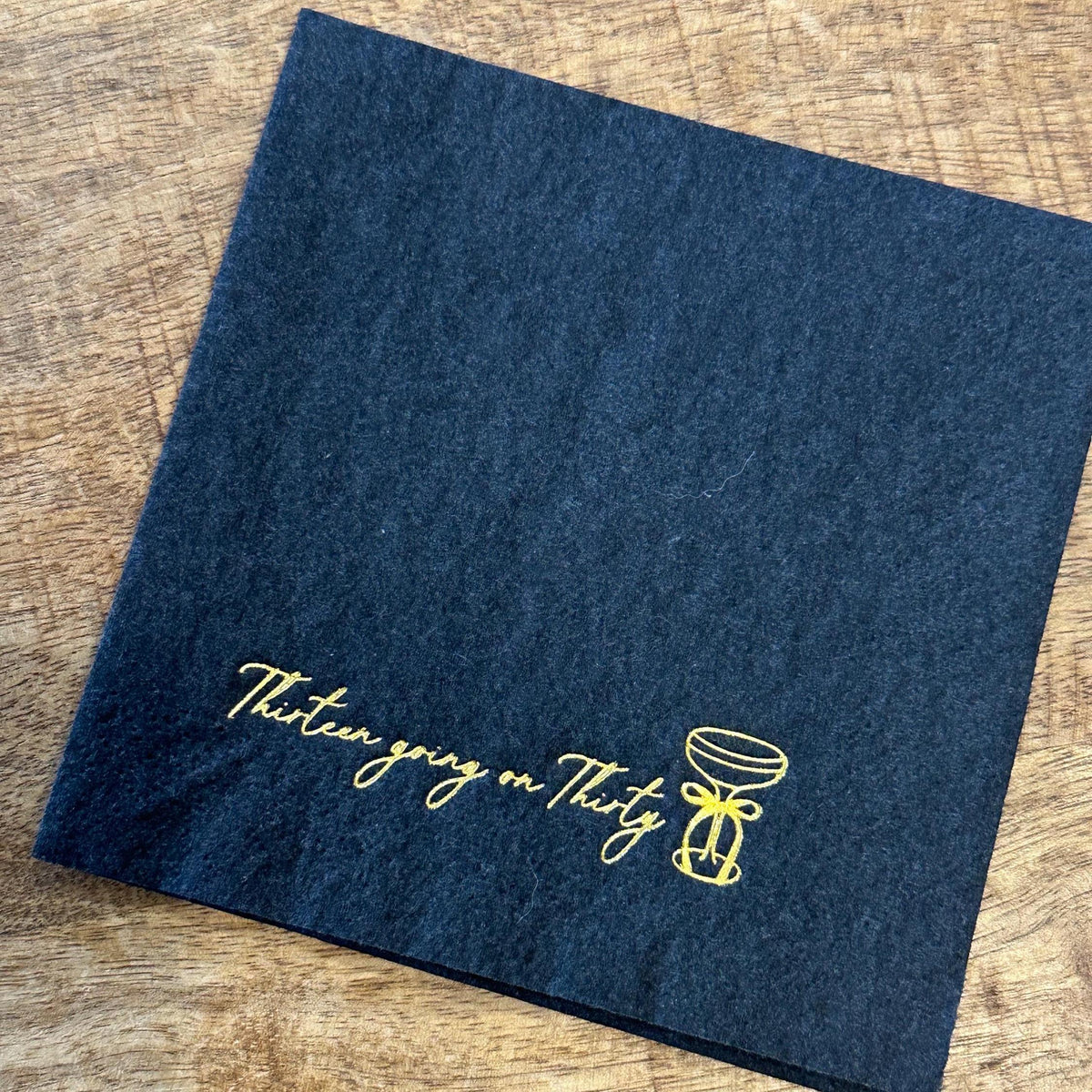A square black paper napkin rests on a wooden table, printed with gold foil text stating Thirteen going on Thirty next to a martini glass illustration.