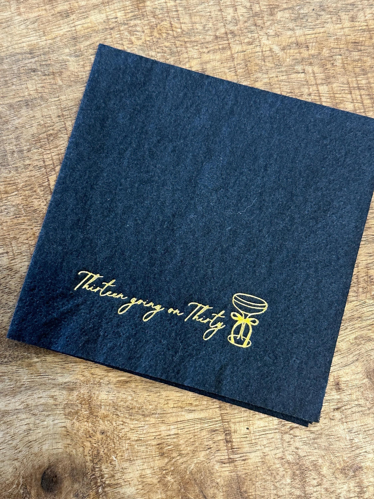 A dark charcoal square paper napkin is placed on a wood grain background, decorated with gold foil script reading Thirteen going on Thirty and a martini glass.