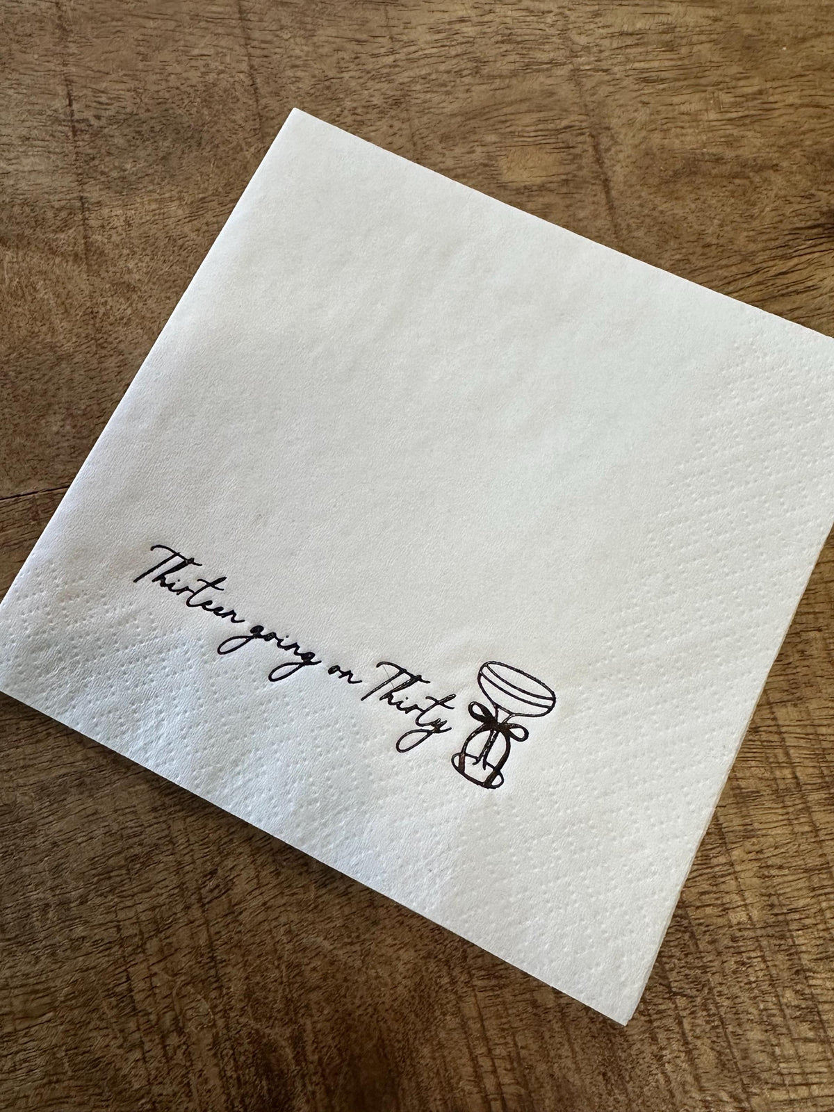 A square white paper napkin is positioned on a wooden surface, displaying black text that says Thirteen going on Thirty alongside a martini glass drawing.