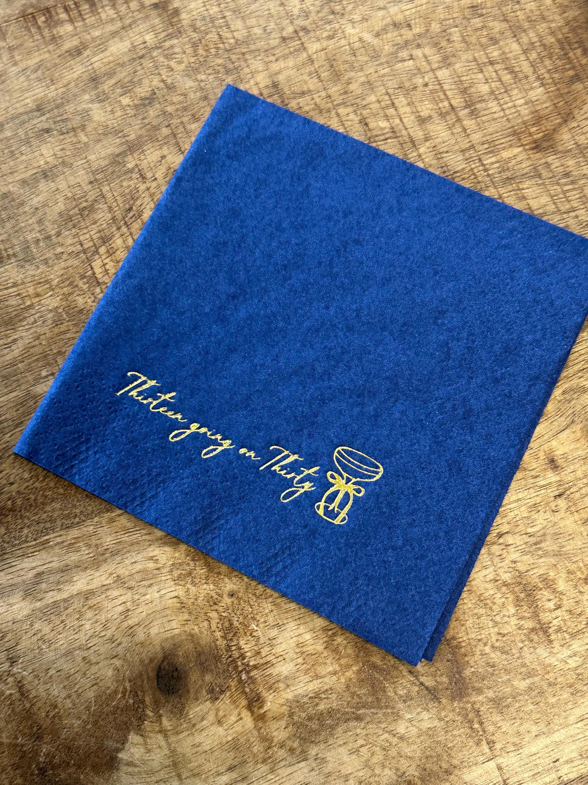 A blue folded paper napkin on a wooden surface, featuring a gold foil-stamped cocktail glass and the text Thirteen going on Thirty.