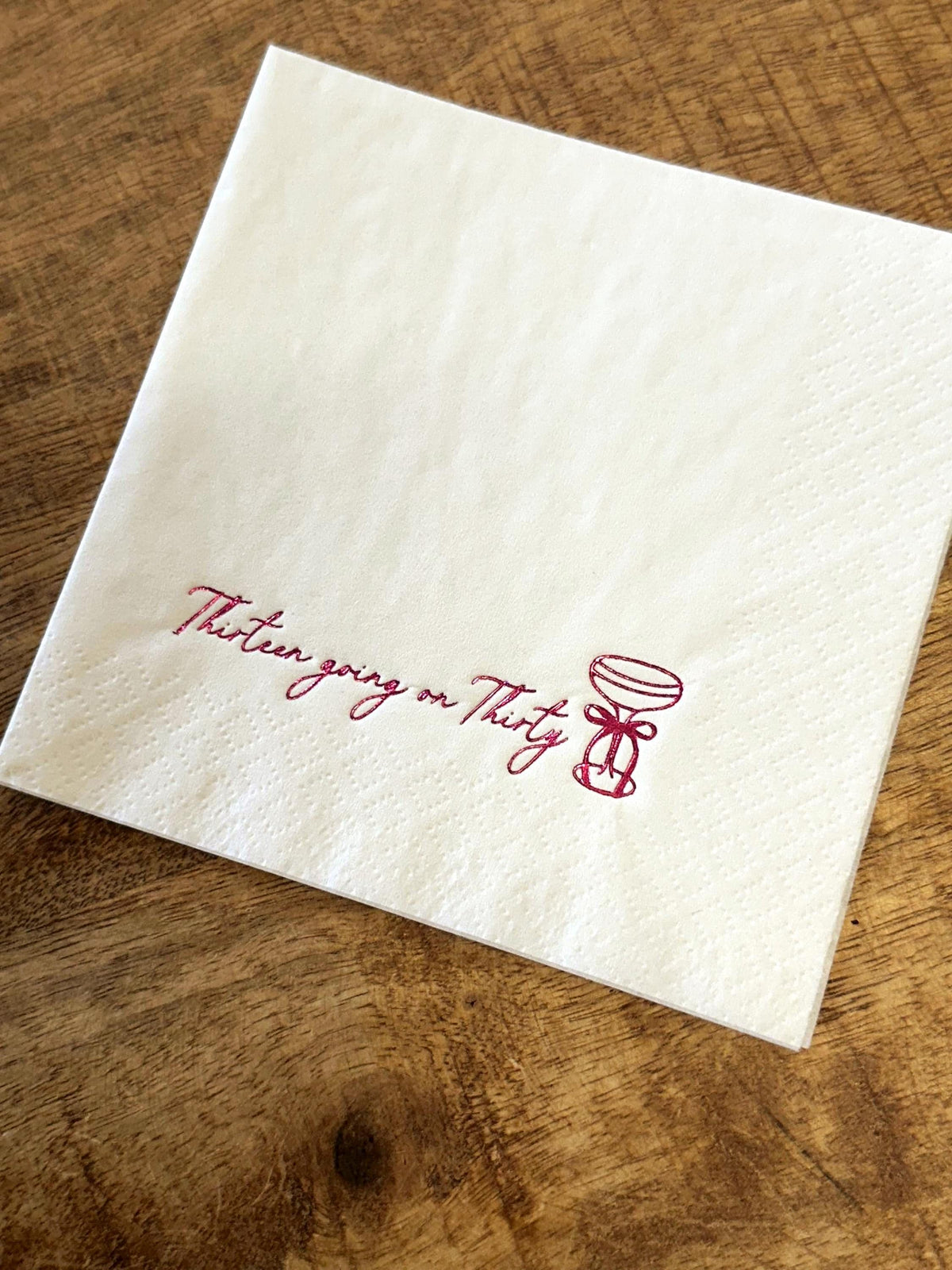 A white folded paper napkin on a wooden surface, featuring a dark pink foil-stamped cocktail glass and the text Thirteen going on Thirty.