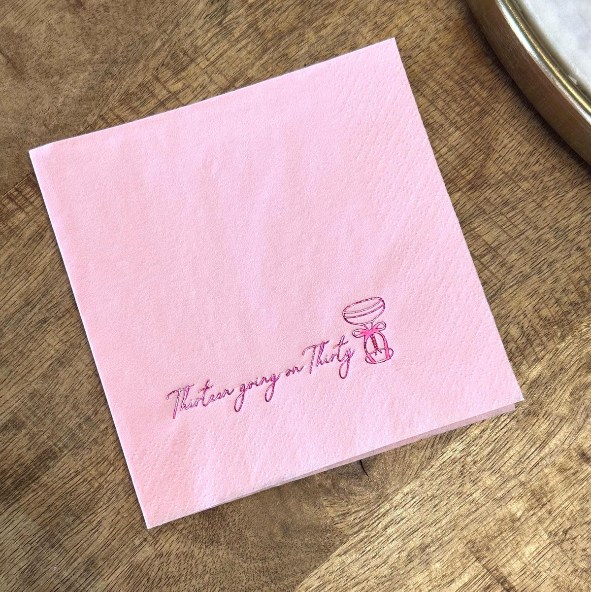A light pink folded paper napkin on a wooden surface, featuring a pink foil-stamped cocktail glass and the text Thirteen going on Thirty.