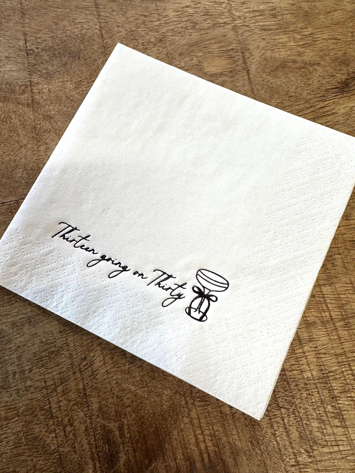 A white folded paper napkin on a wooden surface, featuring a black foil-stamped cocktail glass and the text Thirteen going on Thirty.