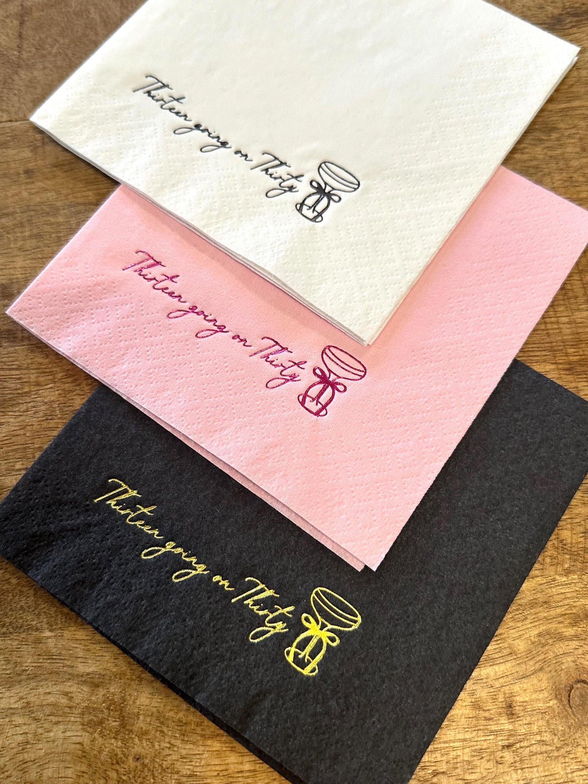 Three folded paper napkins in white, pink, and black stacked at an angle on a wooden surface, printed with the text Thirteen going on Thirty and a cocktail glass.