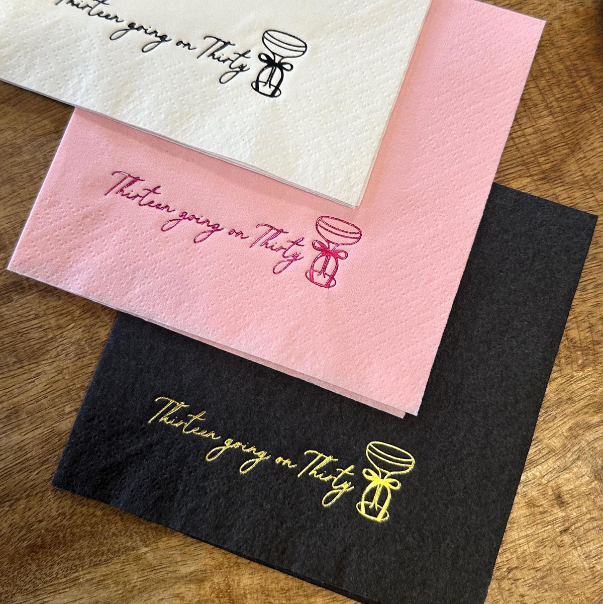Three folded paper napkins in white, pink, and black arranged diagonally on a wooden surface, each printed with the text Thirteen going on Thirty and a cocktail glass graphic.
