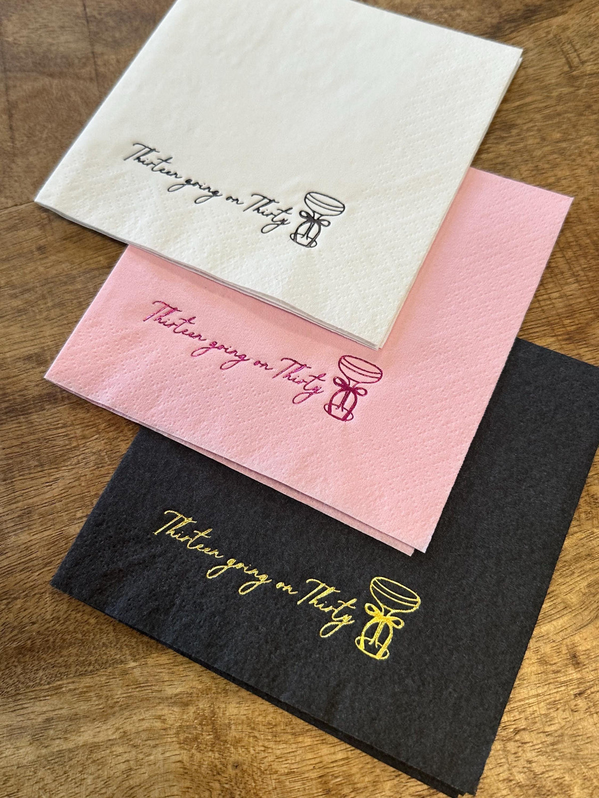 Three folded paper napkins in white, pink, and black stacked on a wooden surface, each featuring foil stamping of the text Thirteen going on Thirty and a cocktail glass.