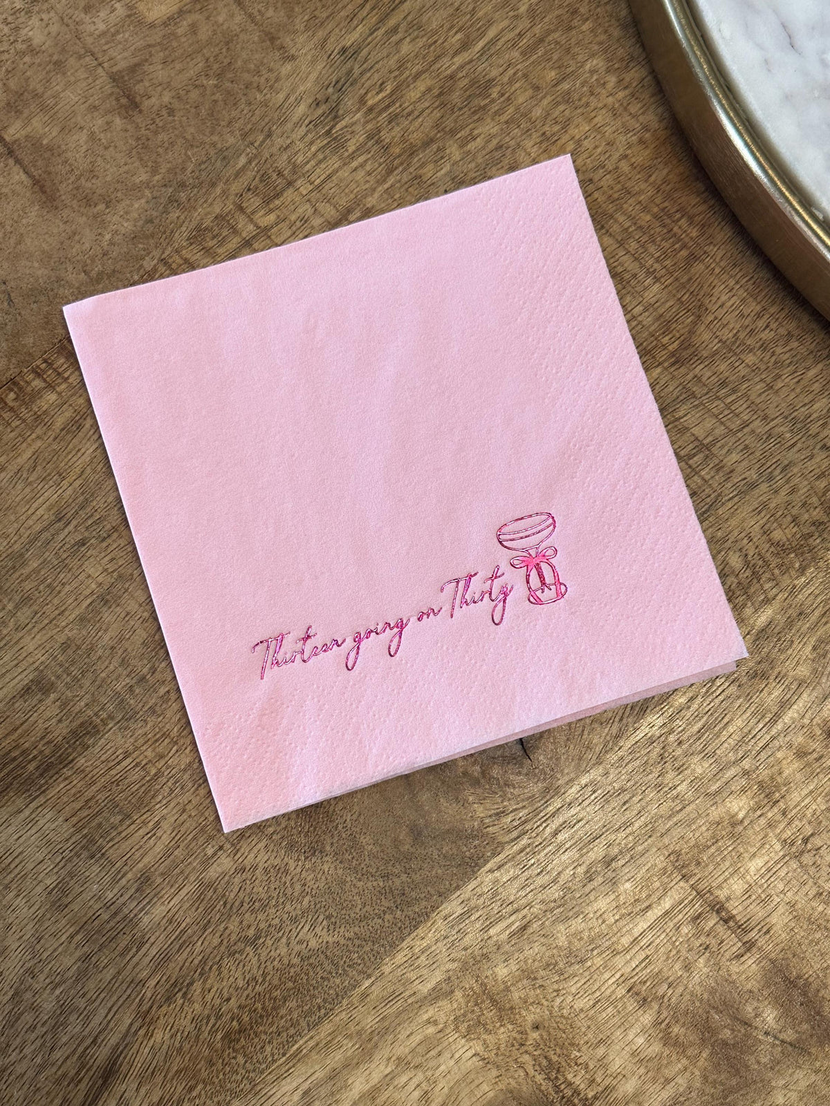 A light pink folded paper napkin on a wooden surface, featuring a dark pink foil-stamped cocktail glass and the text Thirteen going on Thirty.