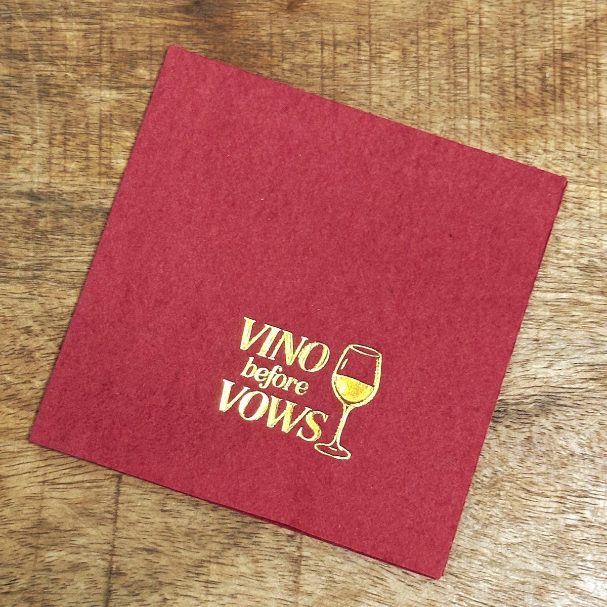 A square burgundy napkin rests on a wooden surface, featuring gold foil text that reads Vino before Vows next to a wine glass.