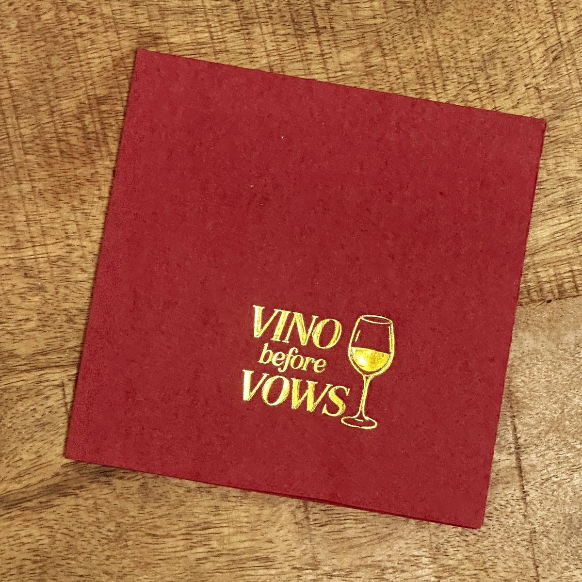 A single deep red cocktail napkin rests on a wooden table, featuring gold foil-stamped text that reads Vino before Vows alongside a wine glass graphic.