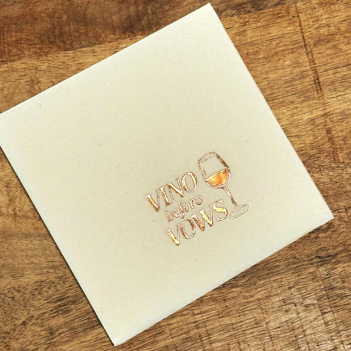 A cream-colored cocktail napkin is displayed on a wooden tabletop, stamped with metallic foil text reading Vino before Vows and a wine glass.