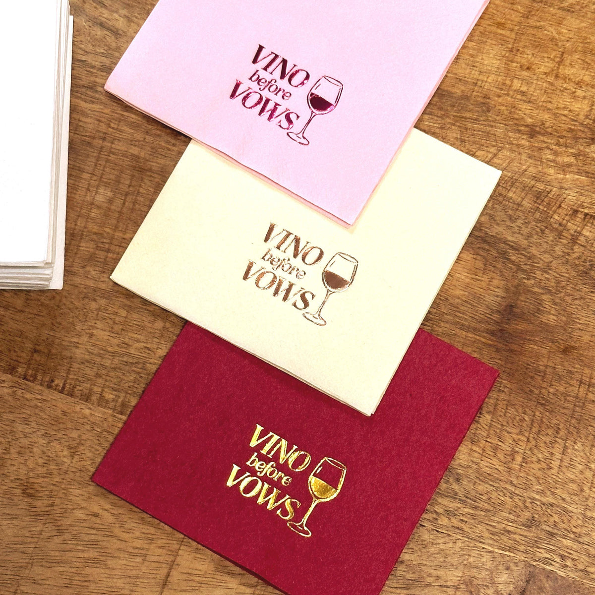 A stack of white napkins sits beside three folded cocktail napkins in pink, cream, and deep red, all printed with Vino before Vows and a wine glass design.