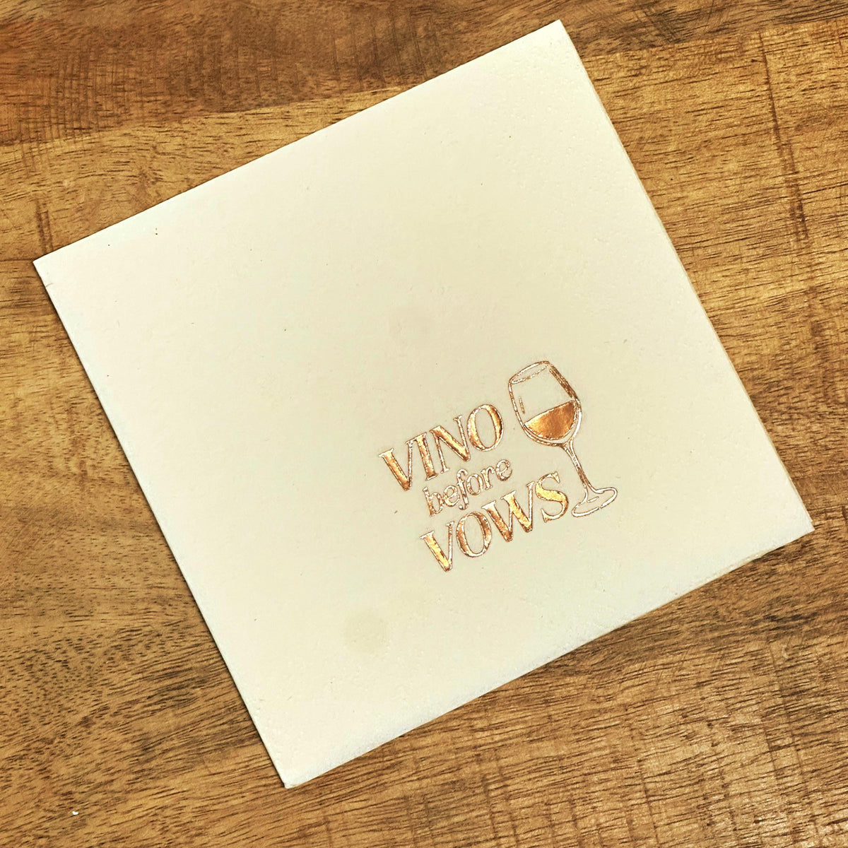 A square white napkin on a wooden table features rose gold foil text reading Vino before Vows alongside a wine glass graphic.