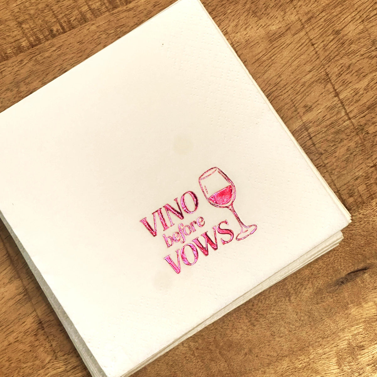 A stack of white square napkins sits on a wooden surface, stamped with pink foil text that reads Vino before Vows and a wine glass.