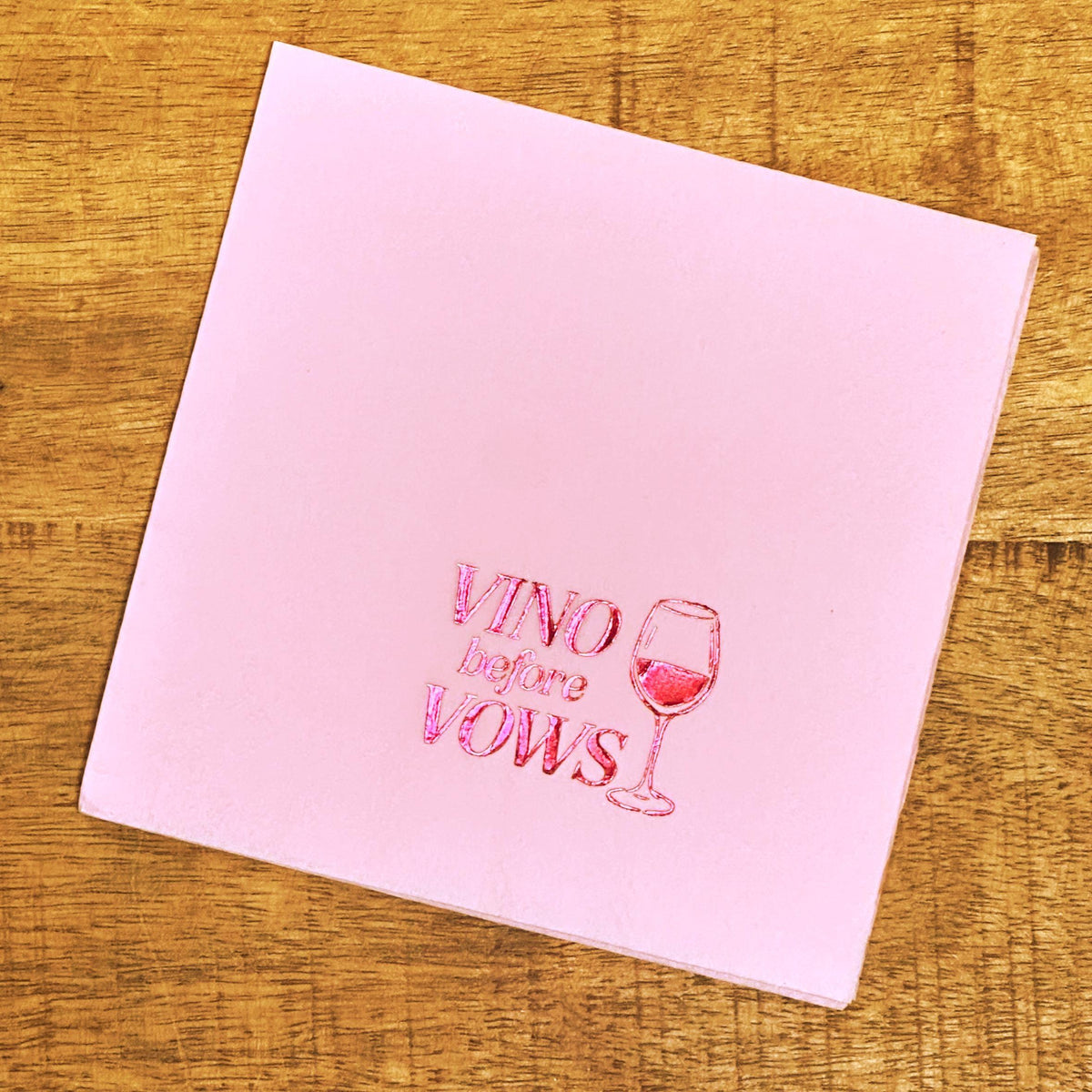 A square light pink napkin on a wooden background displays pink foil stamping with the text Vino before Vows and a wine glass.
