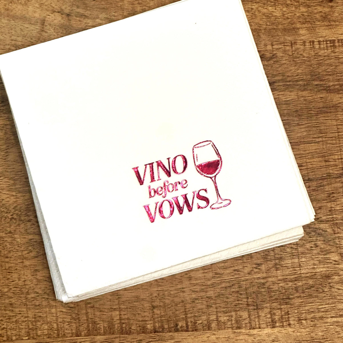 A stack of white square napkins on a wooden table features red foil text and a wine glass illustration.