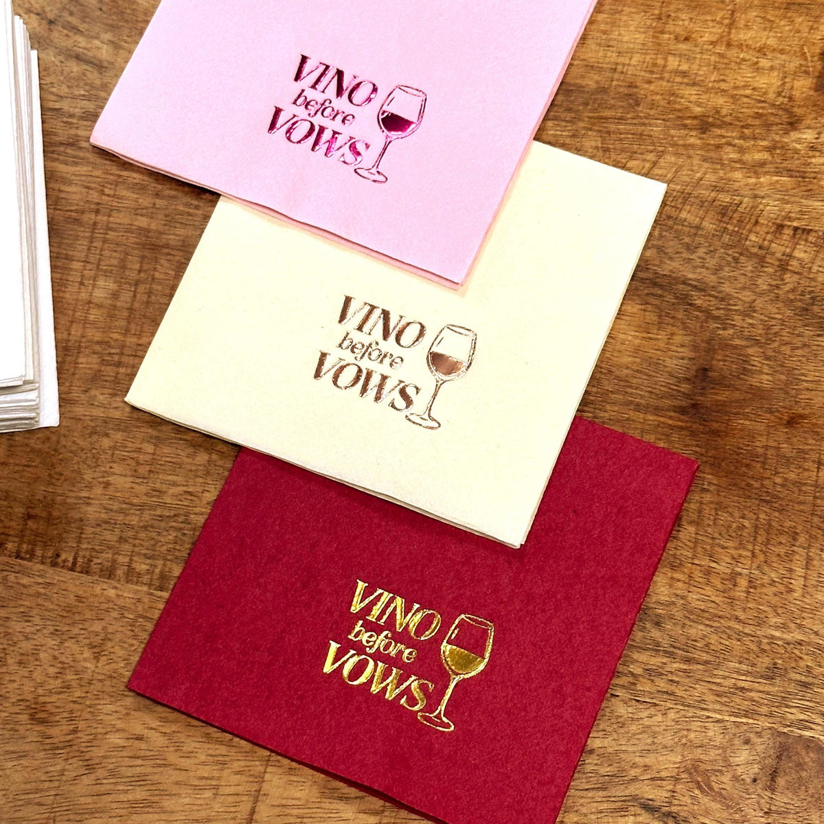 Three folded napkins in pink, cream, and burgundy are arranged diagonally on a wooden table, each featuring foil-stamped text and a wine glass graphic.
