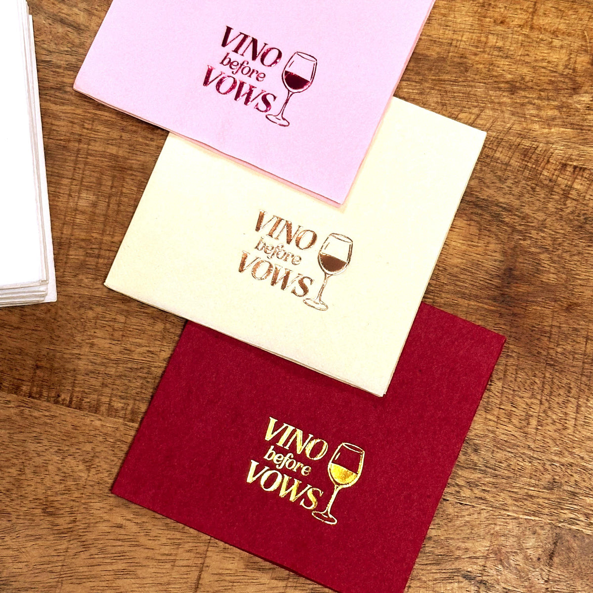 Three folded napkins in light pink, cream, and burgundy lie diagonally on a wooden surface, each featuring foil text and a wine glass graphic.