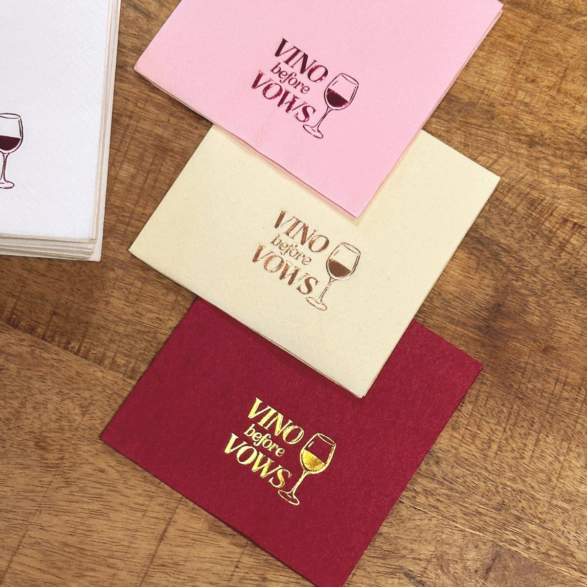 A stack of white napkins rests beside a diagonal arrangement of pink, cream, and burgundy napkins on a wooden table, each featuring foil stamping.