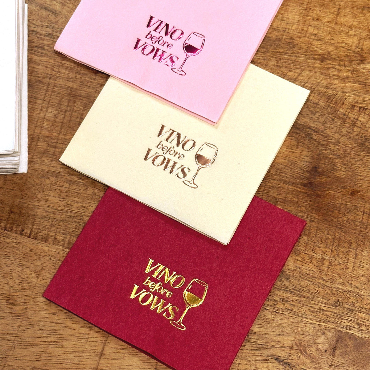 Three folded cocktail napkins in pink, ivory, and deep red are stacked on a wooden surface, each foil-stamped with the text Vino before Vows and a wine glass icon.