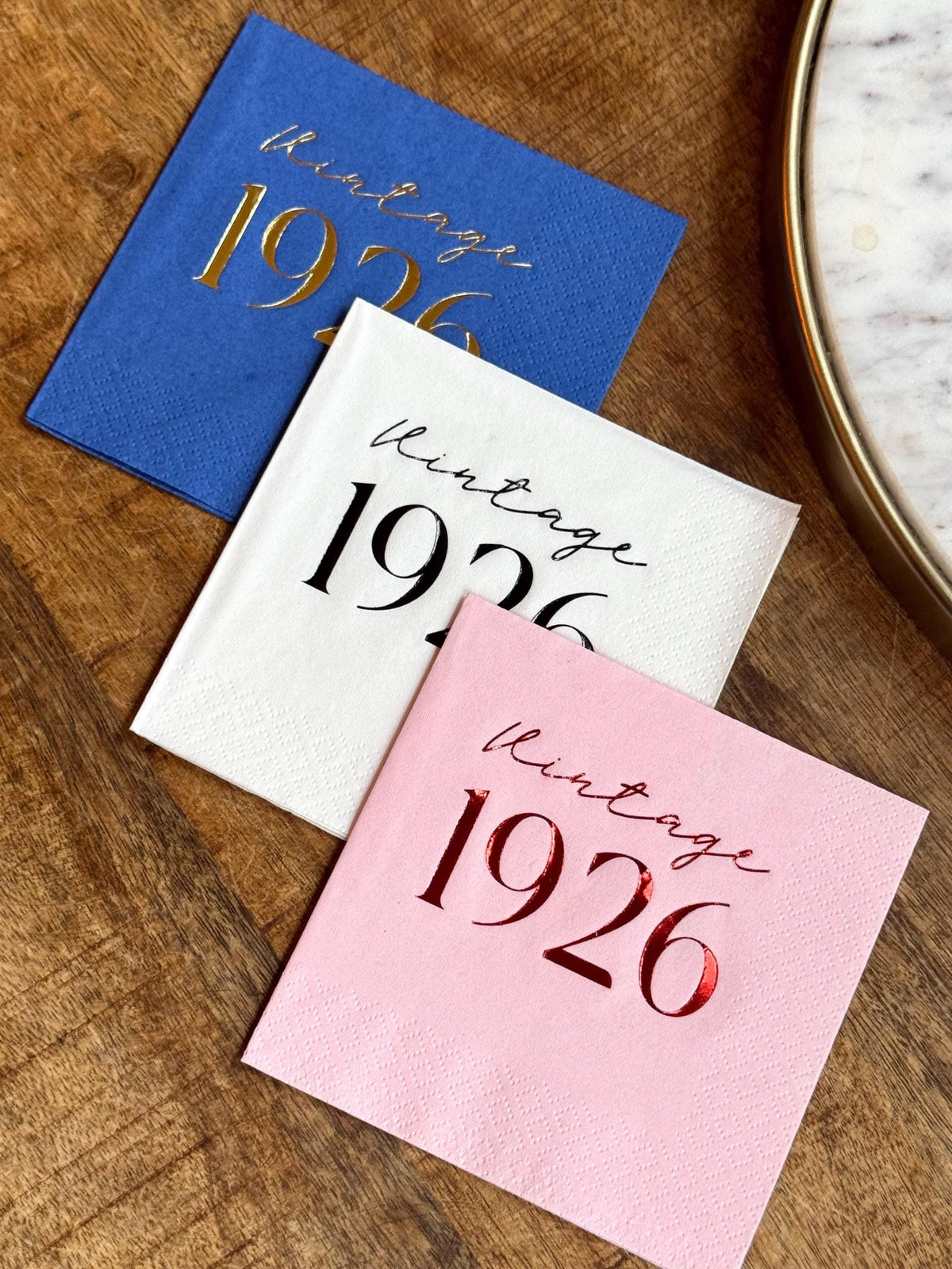 Three folded cocktail napkins in blue, white, and pink are arranged diagonally on a wooden surface, each custom stamped with Vintage 1926 in metallic foil.