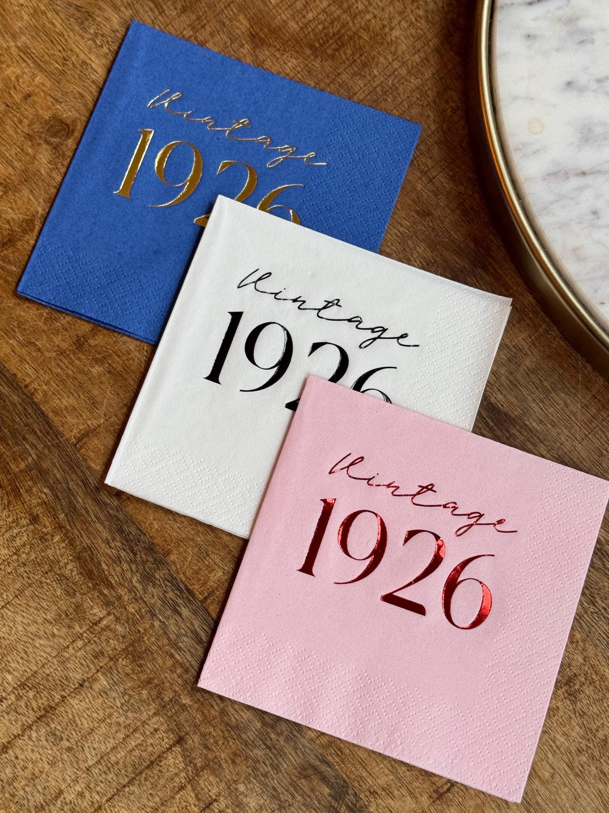 Stack of blue, white, and pink cocktail napkins with Vintage 1926 text, placed on a wooden tabletop.