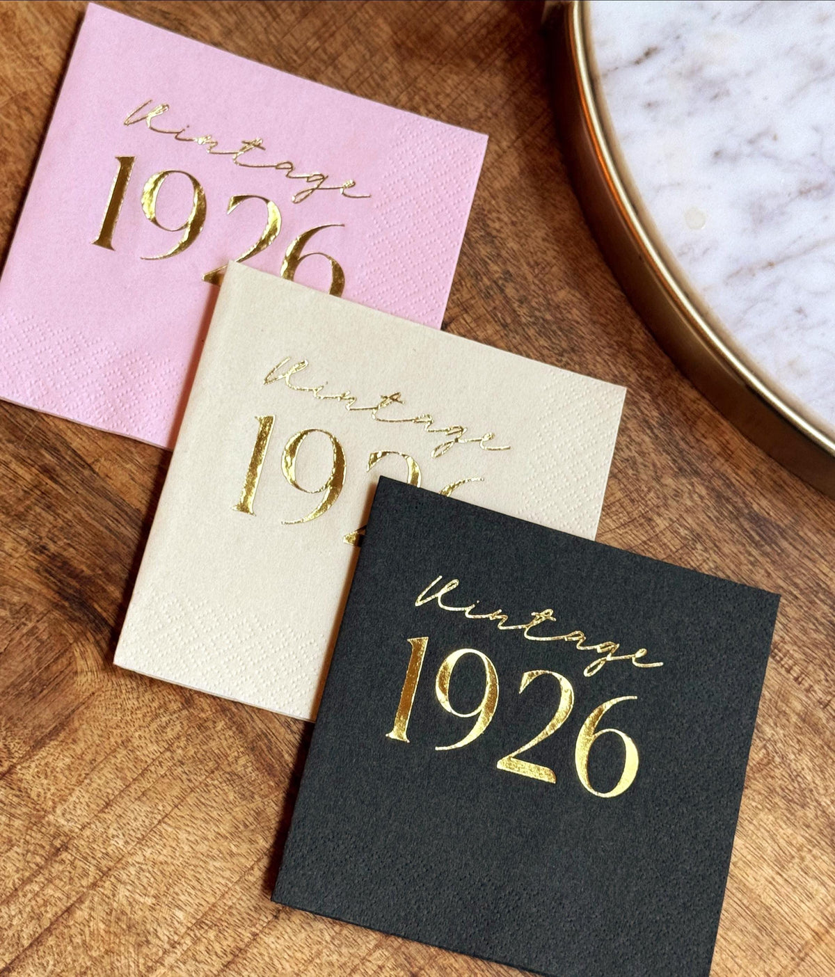Three stacked cocktail napkins in pink, ivory, and black with metallic gold Vintage 1926 foil stamping on a wooden surface.