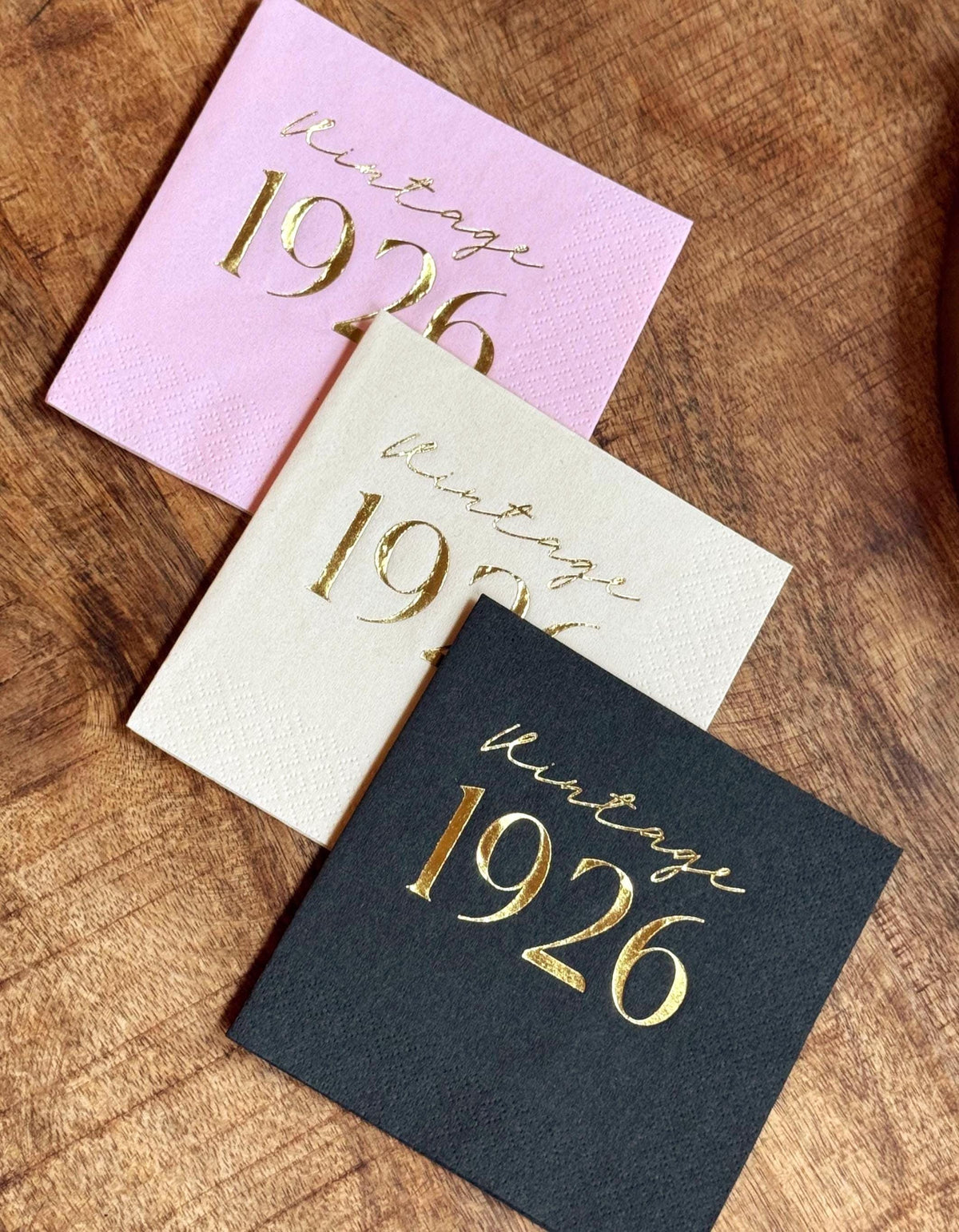Pink, cream, and black napkins featuring gold foil Vintage 1926 text arranged diagonally on a wooden table.