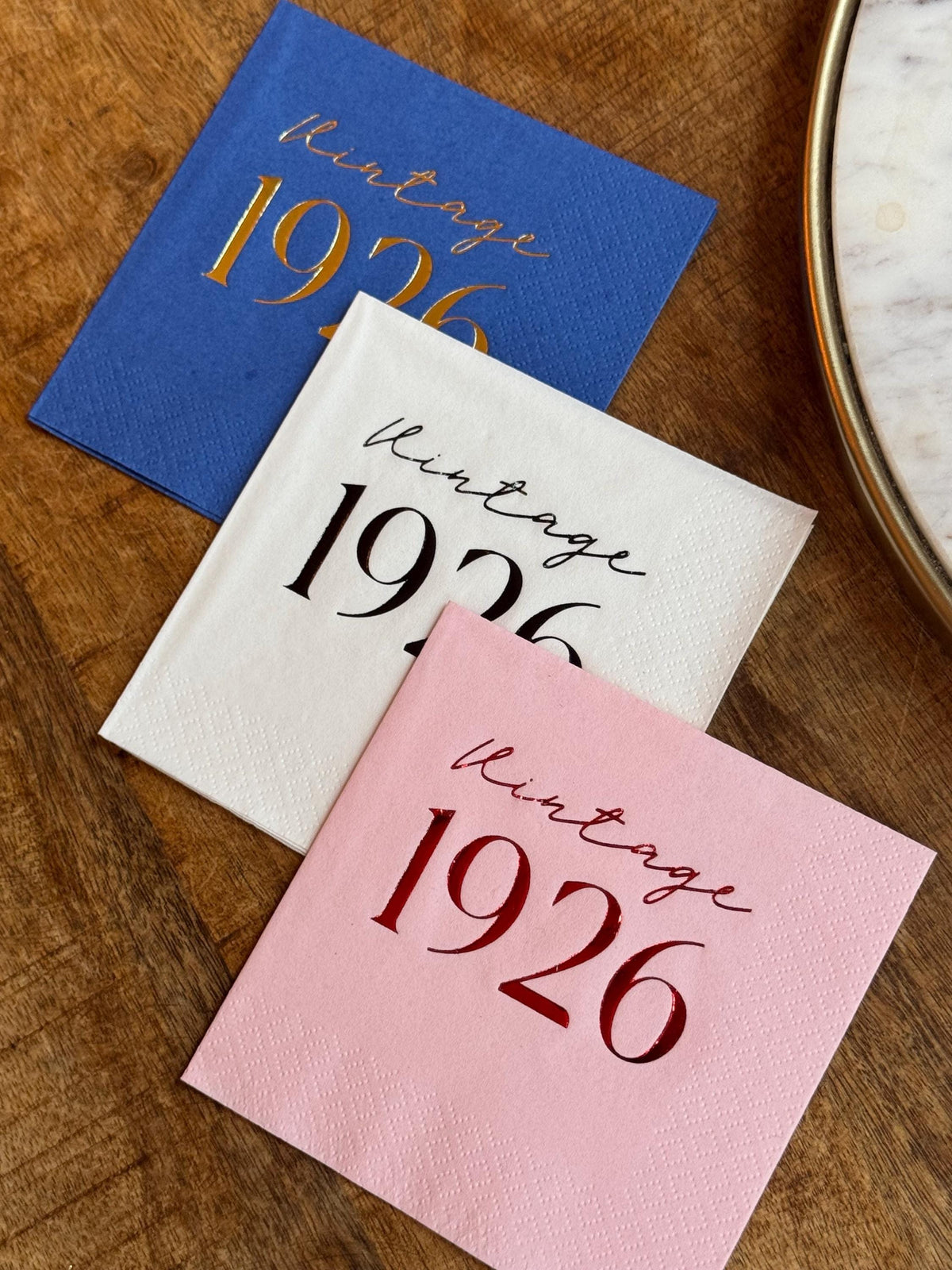 Blue, white, and pink printed cocktail napkins featuring the text Vintage 1926, displayed on a wooden surface next to a tray.